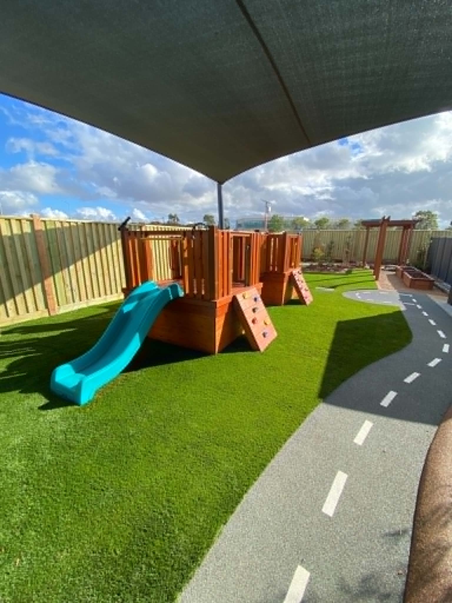 A vibrant early learning outdoor space with synthetic turf, shaded play equipment, soft-fall areas, and a custom roadway path designed for safe, imaginative play.