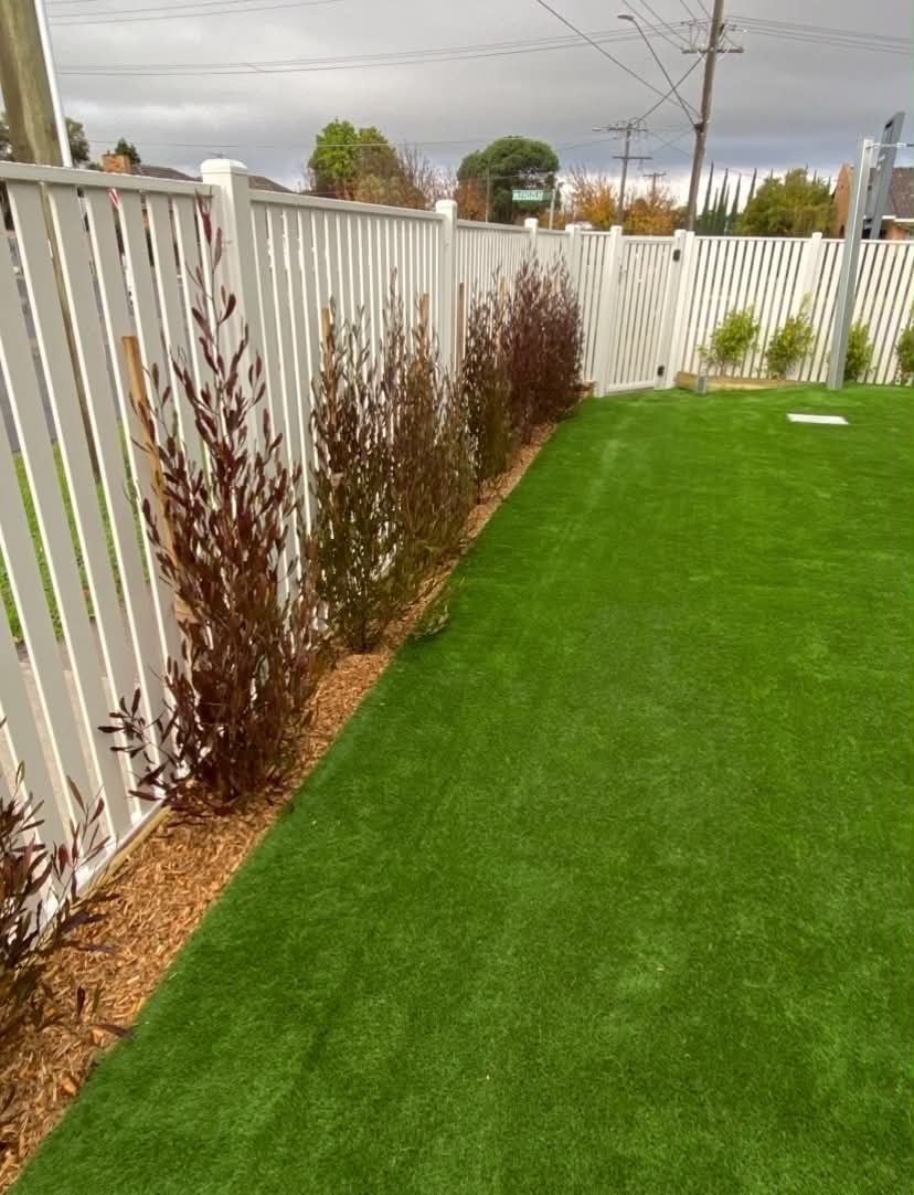A clean and practical childcare outdoor space with synthetic turf, secure boundary fencing, mulch garden beds, and screening plants designed to create a safe, low-maintenance play environment.