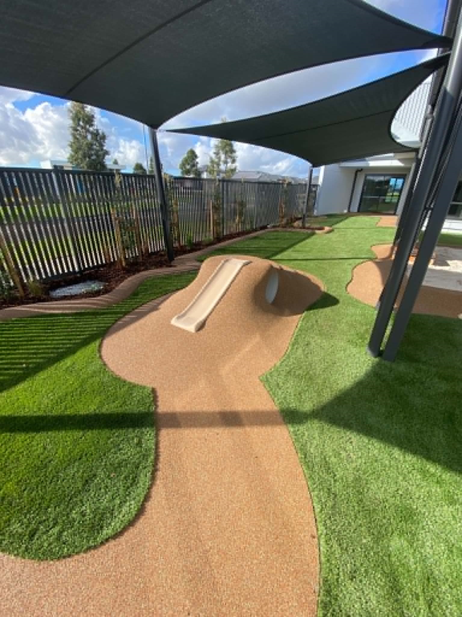 A professionally designed childcare outdoor area with soft-fall pathways, synthetic turf, shaded play zones, landscaped planting, and a built-in slide mound to encourage safe, active exploration.