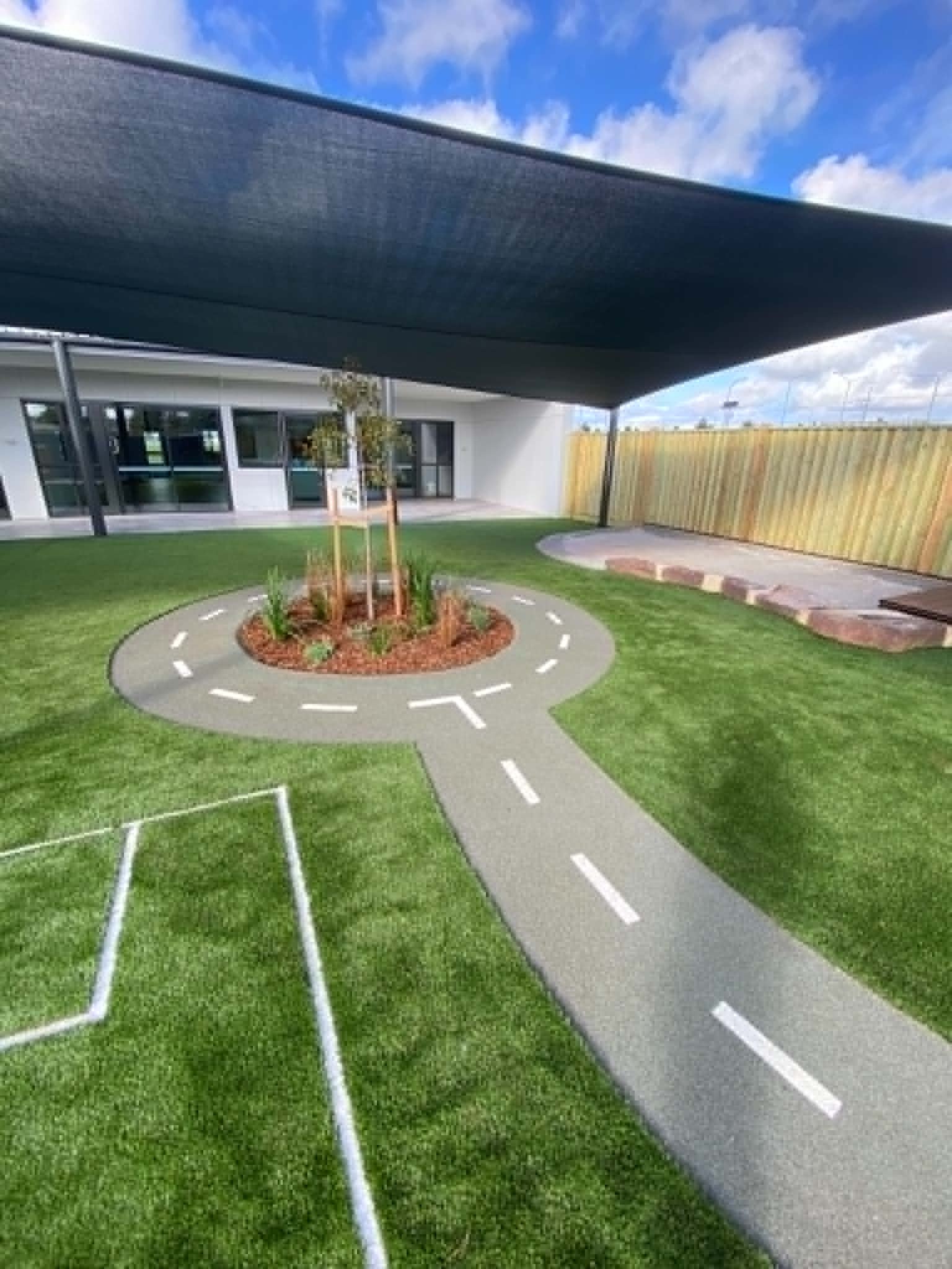 A professionally landscaped childcare play area with soft synthetic turf, shaded outdoor zones, sensory garden planting, and a custom roadway path designed for safe, imaginative learning.