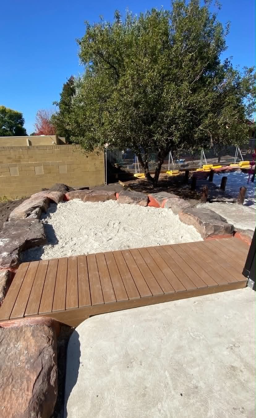A natural childcare play space with sand, timber decking, rock edging, and shaded outdoor areas designed to encourage sensory exploration and imaginative play.