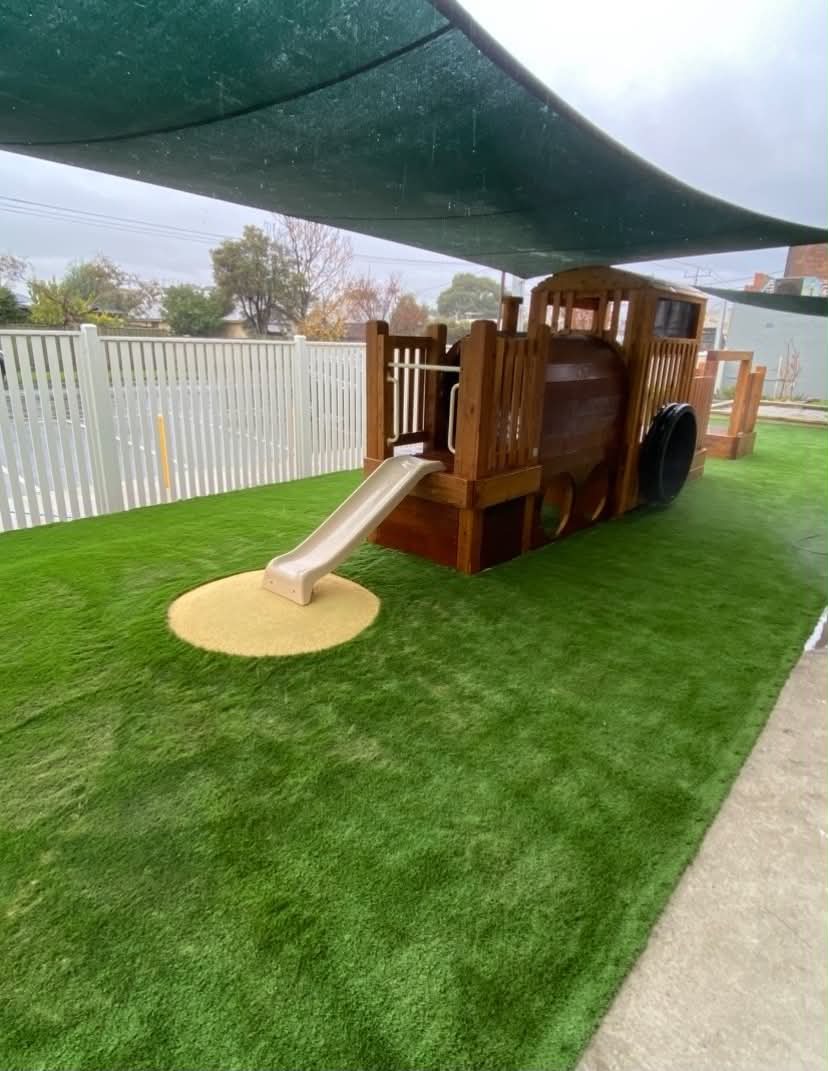 A safe and inviting early learning play space with synthetic turf, shaded timber play equipment, soft-fall surfacing, and room for active outdoor movement.