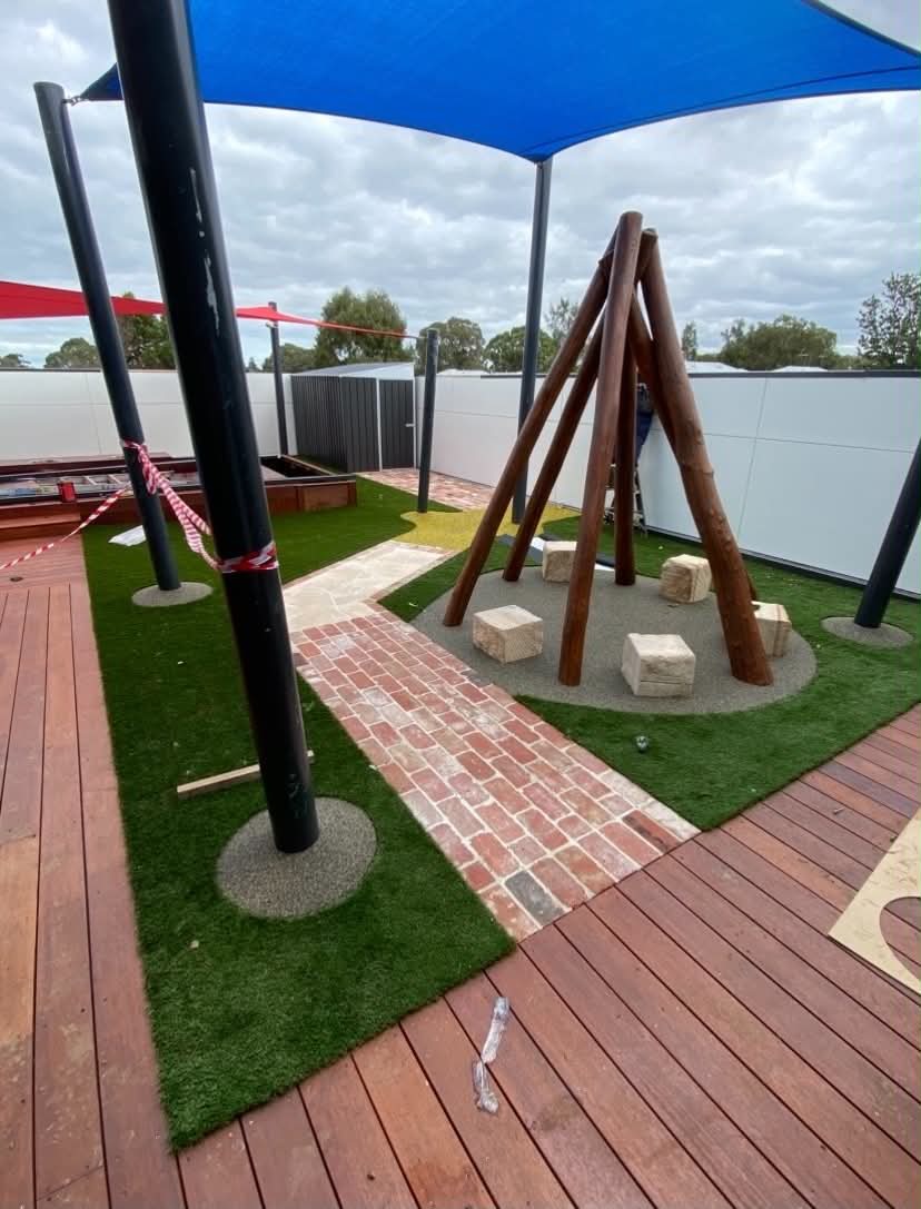 A thoughtfully designed early learning play area with shade sails, synthetic turf, timber decking, soft-fall surfacing, and natural climbing features for active, imaginative play.