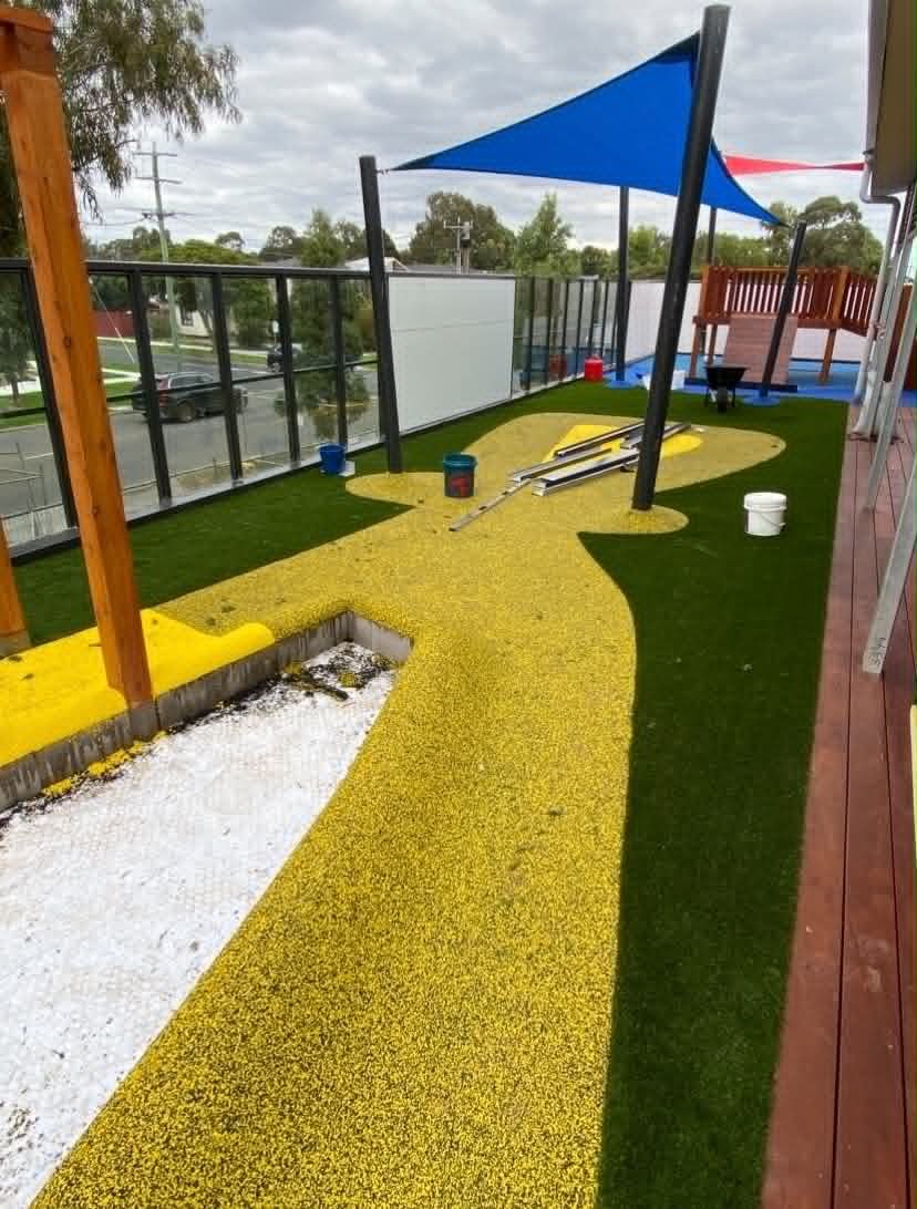 A vibrant childcare outdoor area with soft-fall surfacing, synthetic turf, shaded play spaces, timber edging, and open zones designed for safe, active learning.