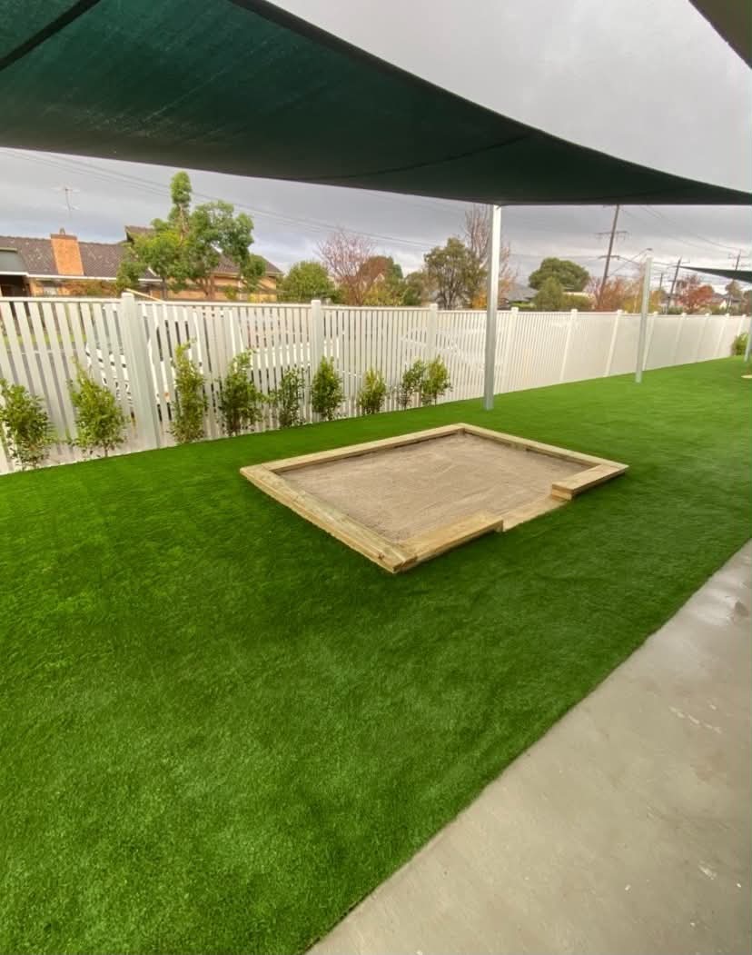 A clean and functional early learning outdoor area with synthetic turf, shaded play space, privacy planting, and a timber sandpit for safe, sensory outdoor learning.