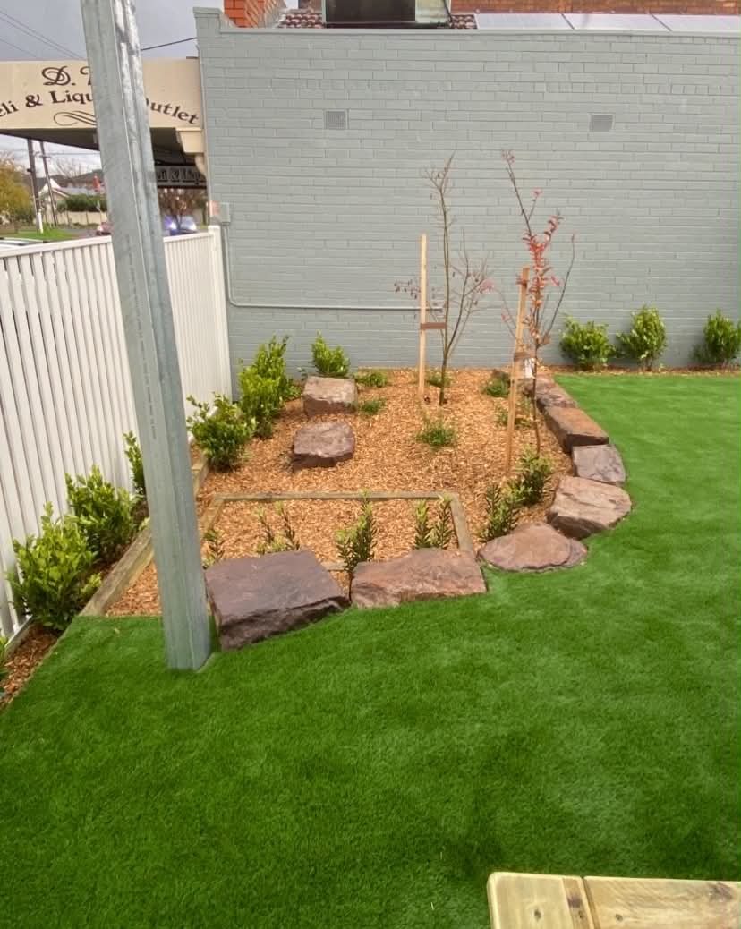 A child-friendly sensory garden space with synthetic turf, natural rock edging, soft mulch, young trees, and planting designed to create a calm outdoor learning area.